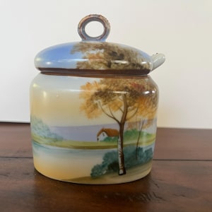 May include: A ceramic jar with a lid, featuring a landscape scene. The jar has a cylindrical shape with a lid that has a handle. The landscape design includes a house, trees, and a body of water, painted in shades of yellow, brown, blue, and green.