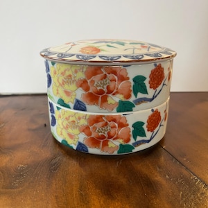 May include: A two-tiered ceramic container with a lid. The container is decorated with large orange and yellow floral designs, green leaves, and red berries. The lid features similar floral patterns and a blue border. The container is on a wooden surface.