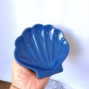 May include: A glossy, deep blue ceramic dish shaped like a scallop shell. The dish features a smooth, concave surface with distinct, radiating ridges that extend from the base to the scalloped outer edge, creating a functional, shell-like form.
