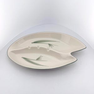 May include: An off-white ceramic dish shaped like a leaf with a divided interior. The surface features a painted green botanical motif of slender leaves. The rim is outlined in a thin dark gray line, and the center divider includes two notches.