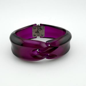 May include: A bold, purple bracelet with a unique, intertwined design. The bracelet is made of a translucent material, with a dark purple hue that fades to a lighter shade. It features a silver-colored clasp.