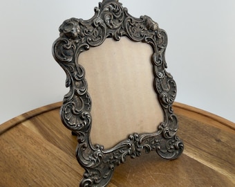 May include: An ornate, dark metallic picture frame with a complex, raised scrollwork and floral relief pattern. The frame features a prominent, pointed top crest and a square-shaped central opening filled with a plain, light tan backing board.