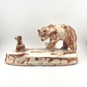 May include: A ceramic bear figurine with a cub. The bear is brown and cream-colored, standing on a cream-colored base with brown marbling. The cub is smaller, brown, and sits on the base. The figurine is a decorative item.