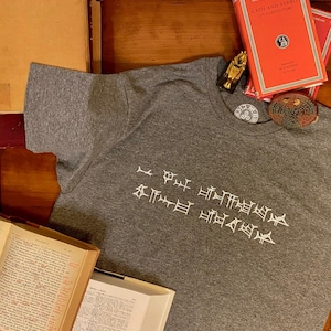 Puede incluir: Una camiseta gris con bordado blanco de una escritura antigua. La escritura dice "Pongo mi esperanza en los dioses".