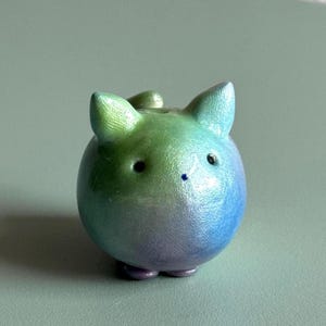 Könnte beinhalten: Eine kleine, kugelförmige Katzenfigur mit spitzen Ohren. Die Oberfläche weist einen glatten, perlmuttartigen Farbverlauf von Grün oben bis hin zu Blau und Violett an der Basis auf. Sie hat zwei winzige schwarze Augen und eine kleine blaue Nase.