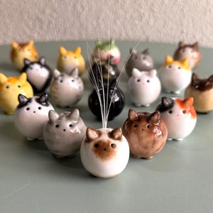 Könnte beinhalten: Eine Sammlung kleiner, kugelförmiger Keramik-Katzenfiguren in verschiedenen Farben und Mustern, darunter Weiß, Braun, Schwarz, Orange und Grau. Jede Figur hat spitze Ohren und ein einfaches Gesicht. Eine zentrale Katze hat dünne, weiße Schnurrhaare.