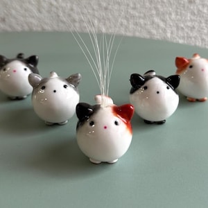 以下が含まれることがあります： 小さな丸いセラミック製の猫の置物のコレクション。各猫は白い体、黒または灰色の耳、小さなピンクの鼻をしています。1匹の猫は細い白い茎の束を持っています。猫は明るい緑色の表面に配置されています。