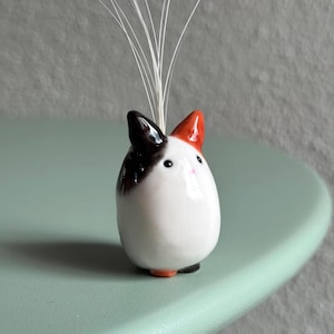 Könnte beinhalten: Eine kleine, katzenförmige Keramikvase mit weißem Körper, schwarzen und orangefarbenen Ohren und einer rosa Nase. Die Vase enthält mehrere dünne, weiße Stiele. Die Vase steht auf einer hellgrünen Oberfläche.