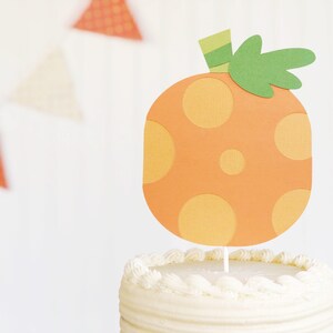 May include: A cake topper shaped like a carrot, with orange and yellow polka dots, green leaves, and a light green stem. The topper is placed on top of a white frosted cake. Orange and white triangle bunting is in the background.