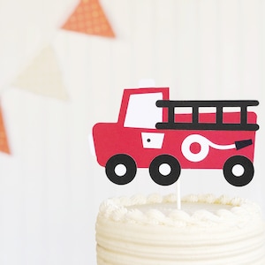 May include: Red and black fire truck cake topper with a white hose and ladder. The topper is on top of a white frosted cake.