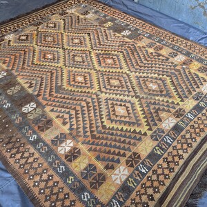 May include: A rectangular rug with a geometric diamond pattern in shades of brown, orange, and yellow. The rug has a fringed edge and a border with repeating geometric shapes. The rug is likely made of woven material.