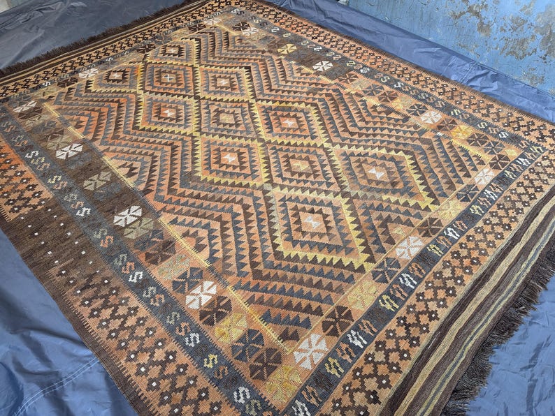 May include: A rectangular rug with a geometric diamond pattern in shades of brown, orange, and yellow. The rug has a fringed edge and a border with repeating geometric shapes. The rug is likely made of woven material.