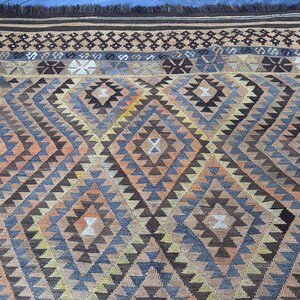 May include: A detailed shot of a hand-woven rug with a geometric pattern. The rug features a diamond design in shades of brown, blue, orange, and beige. The top edge has a dark brown fringe and a decorative border.