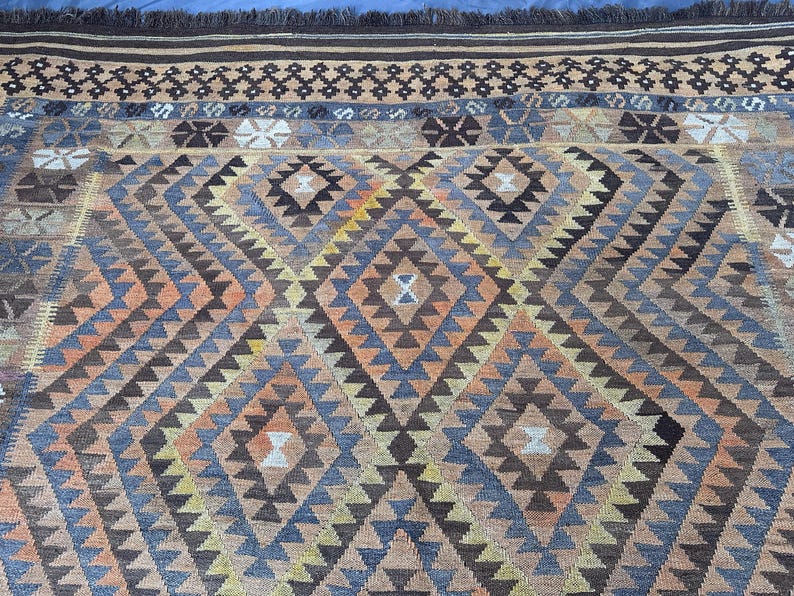 May include: A detailed shot of a hand-woven rug with a geometric pattern. The rug features a diamond design in shades of brown, blue, orange, and beige. The top edge has a dark brown fringe and a decorative border.