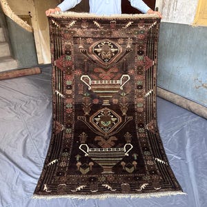 Könnte beinhalten: Ein rechteckiger, dunkelbrauner Teppich mit einem aufwendigen, symmetrischen Muster. Das Design zeigt geometrische Formen, florale Motive und vasenartige Elemente in Braun-, Rot- und Grüntönen. Der Teppich hat einen Fransenrand.