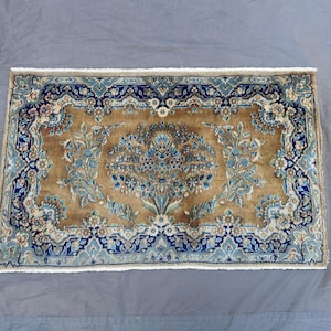 May include: Rectangular rug with a central floral design in shades of blue and beige. The rug features a detailed border in dark blue and light blue, with a beige fringe along the edges. The rug's pattern is symmetrical and intricate.