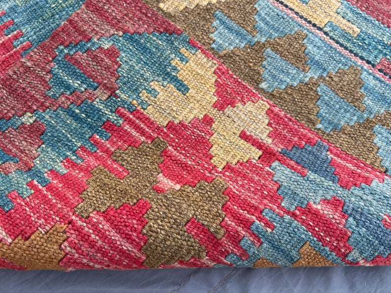 K&ouml;nnte beinhalten: Nahaufnahme eines gewebten Teppichs mit geometrischem Muster. Der Teppich weist eine Kombination aus roten, blauen, beigen und braunen Farben auf. Das Design umfasst Rauten und Zickzackformen, wodurch eine strukturierte Oberfl&auml;che entsteht.