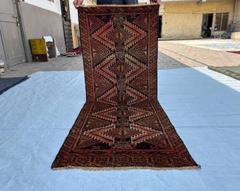 May include: A rectangular runner rug featuring a repeating geometric pattern of diamonds and zigzags in shades of deep red, black, and cream. The rug has a dark border with intricate linear motifs and a short, light-coloured fringe at the bottom edge.