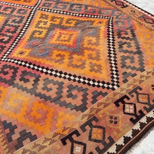 May include: A large, colorful rug with a geometric pattern. The rug features a central diamond design in shades of orange, red, and black, surrounded by a border with repeating motifs. The rug's design is intricate and detailed.