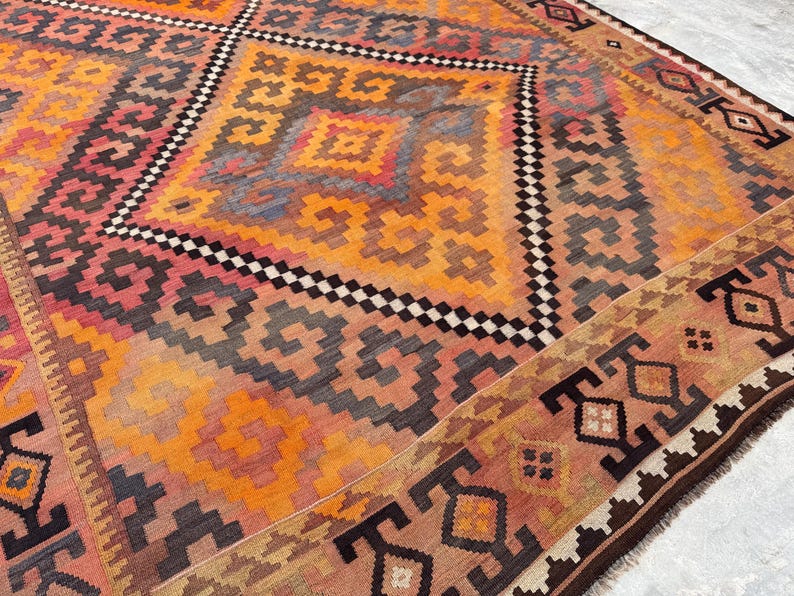 May include: A large, colorful rug with a geometric pattern. The rug features a central diamond design in shades of orange, red, and black, surrounded by a border with repeating motifs. The rug's design is intricate and detailed.
