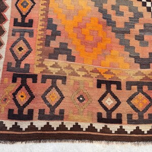 May include: A close-up of a colorful, patterned rug. The rug features geometric designs in shades of orange, pink, brown, and gray. The border has a repeating pattern of diamonds and triangles. The rug is likely made of wool.