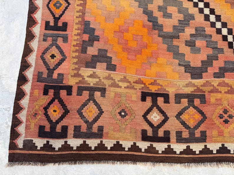 May include: A close-up of a colorful, patterned rug. The rug features geometric designs in shades of orange, pink, brown, and gray. The border has a repeating pattern of diamonds and triangles. The rug is likely made of wool.