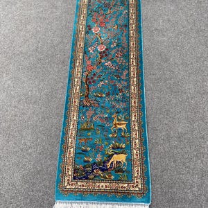 May include: A rectangular runner rug featuring a vibrant turquoise background with a detailed scene of flowering trees, birds, and deer. The rug has a decorative border with intricate patterns and white fringe on both ends of the piece.