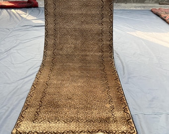 May include: A rectangular brown rug with an intricate geometric pattern and a dark brown border. The rug has a fringe on the shorter ends. The rug is being held up by a person.