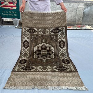 Puede incluir: Una alfombra rectangular con un dise&ntilde;o geom&eacute;trico central en tonos marrones, beige y crema. La alfombra presenta un borde detallado y flecos en los bordes. La alfombra est&aacute; sostenida por una persona.