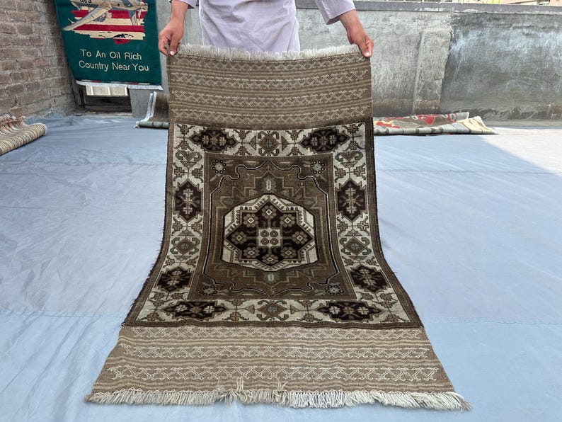 Puede incluir: Una alfombra rectangular con un dise&ntilde;o geom&eacute;trico central en tonos marrones, beige y crema. La alfombra presenta un borde detallado y flecos en los bordes. La alfombra est&aacute; sostenida por una persona.