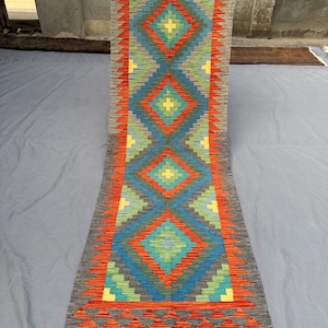 May include: A long, rectangular rug with a geometric pattern. The rug features a central diamond design in shades of blue, orange, green, and yellow, framed by a gray border. The rug has a fringe on the ends.