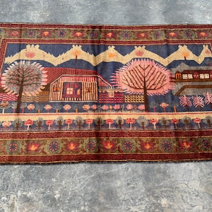 May include: A vintage rug with a colourful design featuring houses, trees, and flowers. The rug has a dark blue background with red, pink, and green accents.