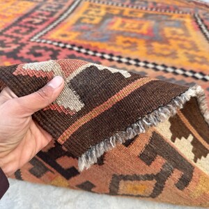 May include: A hand holds a section of a colorful, patterned rug. The rug features a geometric design with shades of orange, brown, and beige. The rug's edge is visible, showing a fringed border and detailed weaving.