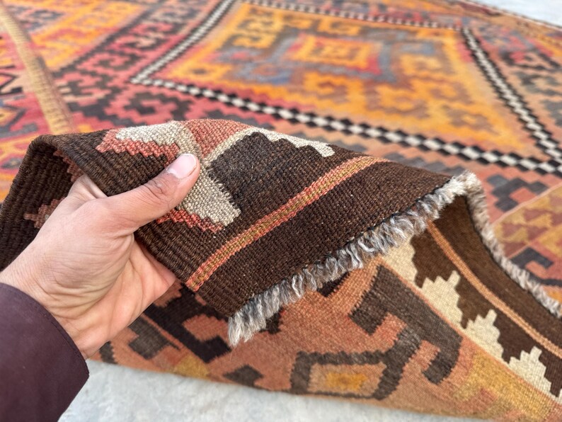 May include: A hand holds a section of a colorful, patterned rug. The rug features a geometric design with shades of orange, brown, and beige. The rug's edge is visible, showing a fringed border and detailed weaving.