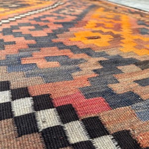 May include: Close-up of a woven rug with a geometric pattern. The rug features a mix of colors, including orange, red, blue, and black. The design includes repeating shapes and a textured surface.