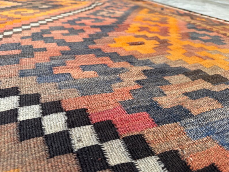 May include: Close-up of a woven rug with a geometric pattern. The rug features a mix of colors, including orange, red, blue, and black. The design includes repeating shapes and a textured surface.
