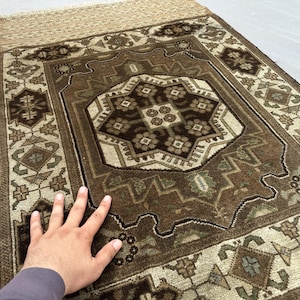 Puede incluir: Una alfombra rectangular marr&oacute;n y beige con un intrincado patr&oacute;n geom&eacute;trico. La alfombra presenta un dise&ntilde;o de medall&oacute;n central con motivos florales y un borde con formas geom&eacute;tricas repetidas. La alfombra tiene un borde con flecos.