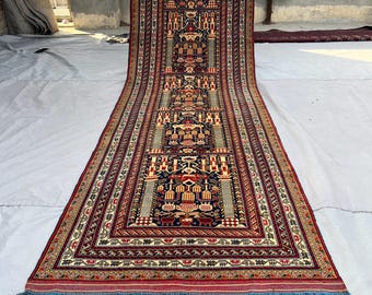 Könnte beinhalten: Ein langer, rechteckiger Teppich mit einem komplizierten geometrischen Muster in Rot-, Blau-, Beige- und Grüntönen. Der Teppich hat ein zentrales Design mit sich wiederholenden Motiven und einen blauen Fransen am Ende. Der Teppich ist wahrscheinlich aus Wolle.