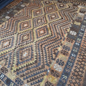 May include: A rectangular rug with a geometric diamond pattern in shades of brown, tan, and blue. The rug features intricate designs and borders with floral and geometric motifs. The overall aesthetic is vintage and handcrafted.