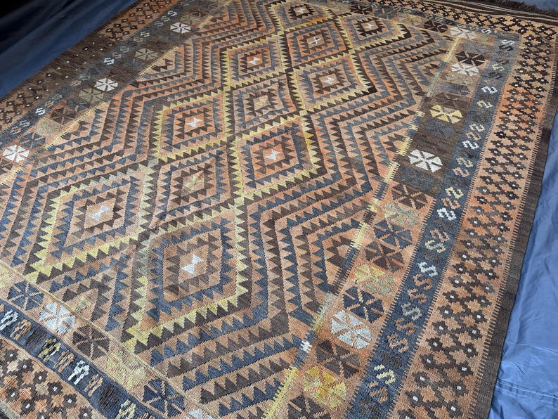 May include: A rectangular rug with a geometric diamond pattern in shades of brown, tan, and blue. The rug features intricate designs and borders with floral and geometric motifs. The overall aesthetic is vintage and handcrafted.