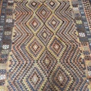 May include: A large, rectangular rug with a geometric diamond pattern in shades of brown, orange, and yellow. The rug has a border with repeating geometric shapes and a blue edge. The rug is likely made of woven material.