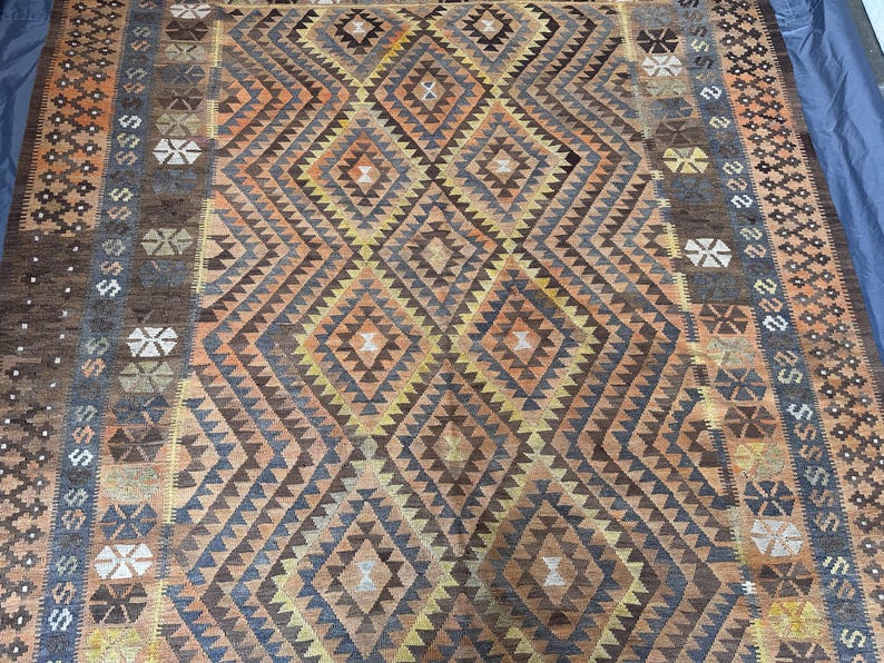 May include: A large, rectangular rug with a geometric diamond pattern in shades of brown, orange, and yellow. The rug has a border with repeating geometric shapes and a blue edge. The rug is likely made of woven material.