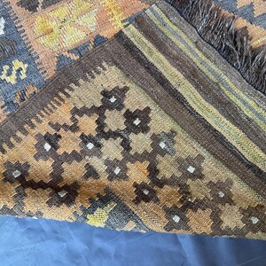 May include: A section of a woven rug with a geometric pattern. The rug features a combination of brown, orange, and blue hues. The edges are finished with a dark brown border and fringe.