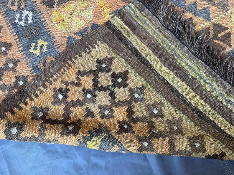 May include: A section of a woven rug with a geometric pattern. The rug features a combination of brown, orange, and blue hues. The edges are finished with a dark brown border and fringe.