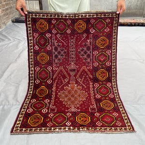 May include: A rectangular, hand-woven rug with a deep red background and intricate geometric patterns in shades of brown, gold, and white. The rug features a central medallion design and a detailed border. The rug is displayed on a light-colored surface.