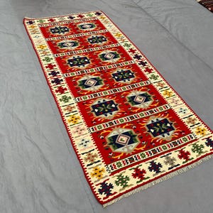 Puede incluir: Una alfombra rectangular tejida a mano con un campo central rojo y patrones geométricos. La alfombra presenta un diseño repetitivo de diamantes y cuadrados en tonos de azul, verde y crema, enmarcado por un borde crema con motivos florales coloridos. La alfombra mide aproximadamente 152 cm de largo.