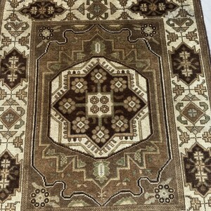 Puede incluir: Una alfombra rectangular con un intrincado patr&oacute;n geom&eacute;trico en tonos marr&oacute;n, beige y verde oliva. El dise&ntilde;o central presenta una forma de diamante marr&oacute;n oscuro con motivos florales, rodeada por un borde beige m&aacute;s claro y bordes detallados.