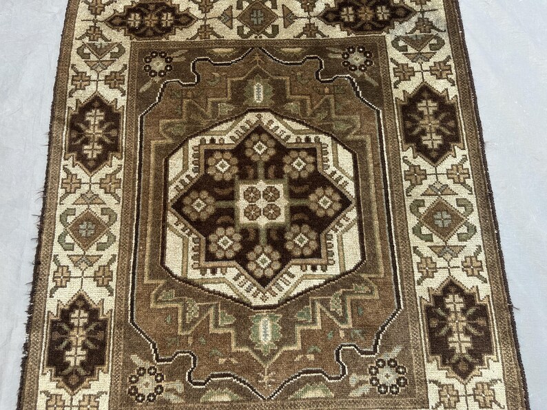 Puede incluir: Una alfombra rectangular con un intrincado patr&oacute;n geom&eacute;trico en tonos marr&oacute;n, beige y verde oliva. El dise&ntilde;o central presenta una forma de diamante marr&oacute;n oscuro con motivos florales, rodeada por un borde beige m&aacute;s claro y bordes detallados.