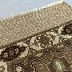 Puede incluir: Primer plano de una secci&oacute;n de una alfombra estampada en tonos marr&oacute;n y beige. La alfombra presenta un dise&ntilde;o geom&eacute;trico repetitivo con detalles en crema. El borde superior tiene flecos. El dise&ntilde;o de la alfombra es detallado e intrincado.