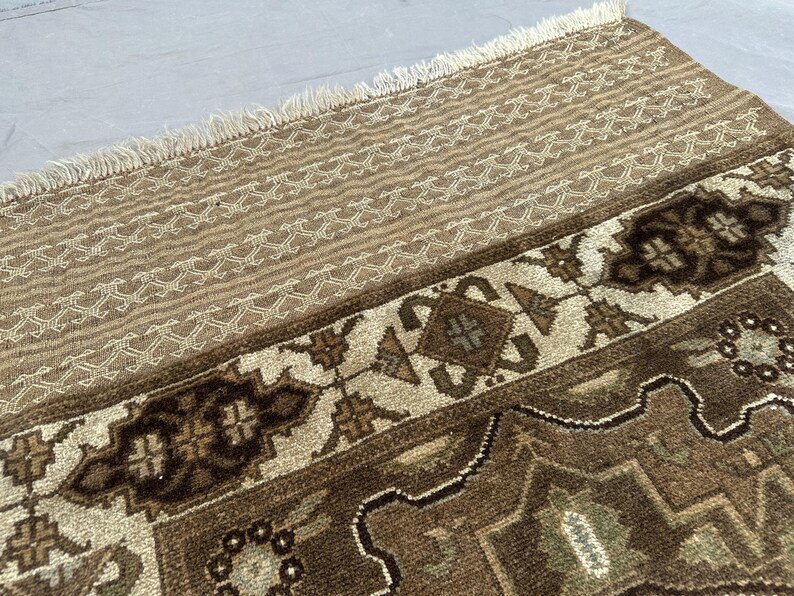 Puede incluir: Primer plano de una secci&oacute;n de una alfombra estampada en tonos marr&oacute;n y beige. La alfombra presenta un dise&ntilde;o geom&eacute;trico repetitivo con detalles en crema. El borde superior tiene flecos. El dise&ntilde;o de la alfombra es detallado e intrincado.